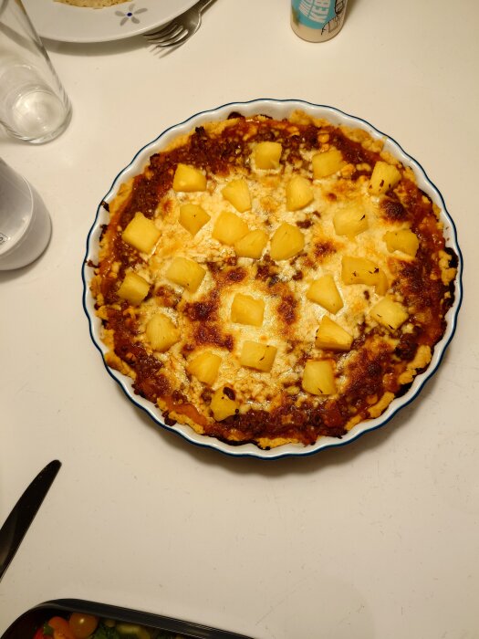 Tacopaj med ananasbitar i en rund form på ett bord, omgiven av bestick och ett glas.