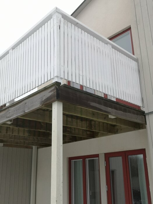 Balkong med vitmålat räcke, stödd av trästolpar på plintar. Stolparna ser ut att bära bjälklaget, utan synlig kontakt med balkongens ram.