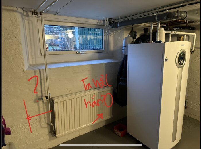 Källare med fönster, radiator och värmepump. Handritade anteckningar frågar var hål ska göras i väggen för installation.