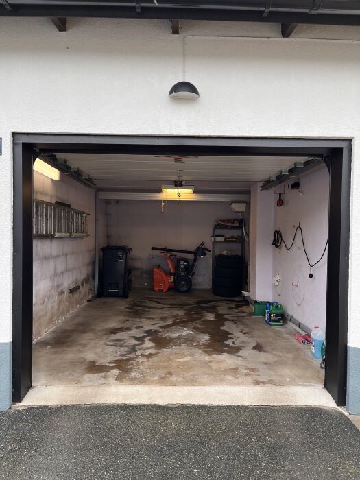 Öppet garage med betonggolv, snöslunga, verktyg och däck. Väggarna är delvis blottade av blåbetong, taklampa och en vägglampa syns.