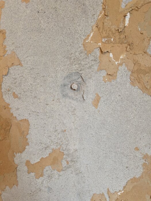 Grå vägg med flagnad tapet och en liten pluggad träbit synlig. Delar av väggen visar slitage och det finns områden med svart material.