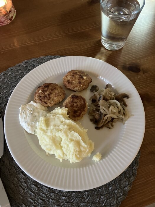 Kycklingfärsbiffar med potatismos, stekt lök och svamp samt citronyoghurt på en vit tallrik, placerad på ett bord med ett glas vatten.