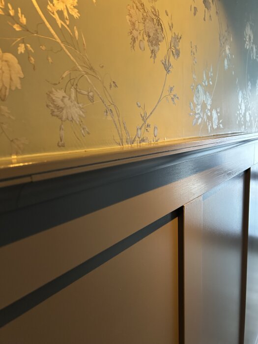 Väggpanel med blommigt mönstrad tapet och trälist i närbild.