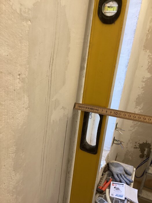 Vägg med byggmaterial och ett vattenpass används för att mäta raka linjer, med tumstock mot väggen. Verktyg och handskar synliga nedanför.