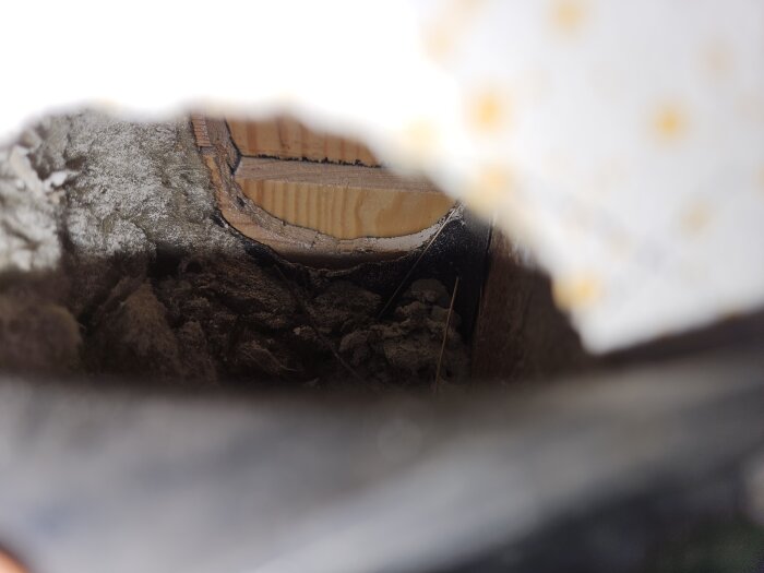 Inuti vägg i badrum, synligt isoleringsmaterial och trä från ett igensatt hål. Eventuell asbest ifrågasätts.