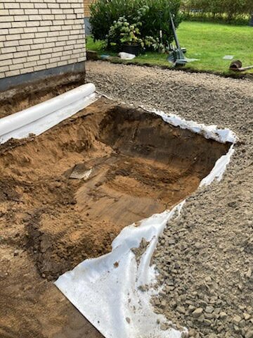 Grävt hål i marken framför garage med synlig jord och grus, samt delvis utrullad markduk runt om.