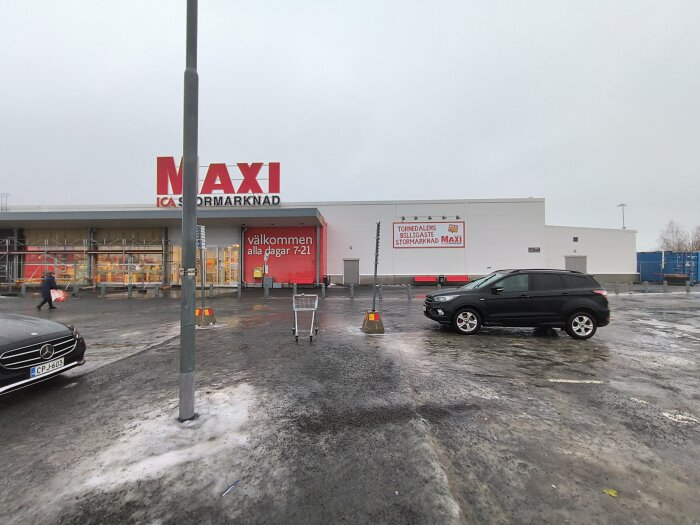 ICA Maxi-butiksfasad med skyltar om öppettider och "Tornedalens billigaste stormarknad". Parkeringsplats med bilar och kundvagn i förgrunden.