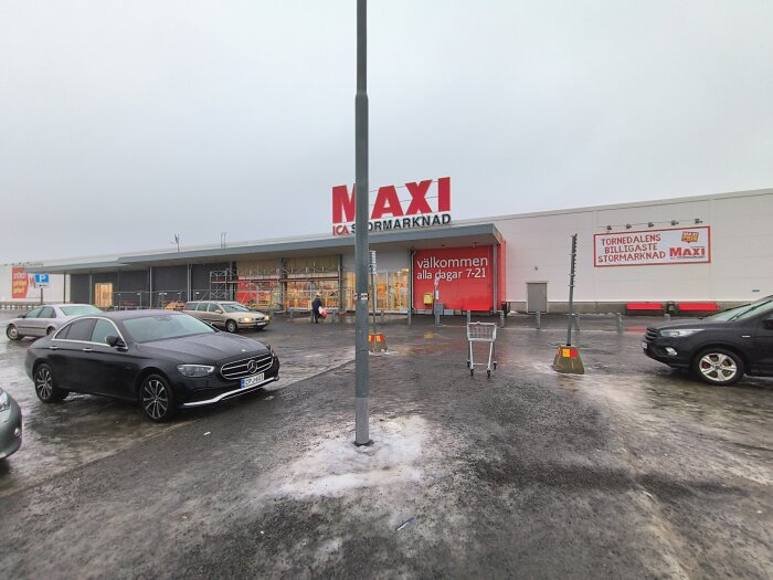 Utanför en ICA Maxi-butik med bilar och kundvagnar på parkeringsplatsen, butiksskylt och entré synliga i bakgrunden.