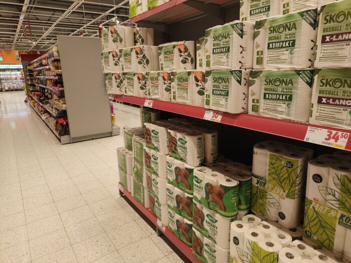 Hyllor med toalettpapper och hushållspapper inne på en Ica Maxi-butik med produkter märkta "Skona" och "Serla". Interiördelen av butiken.