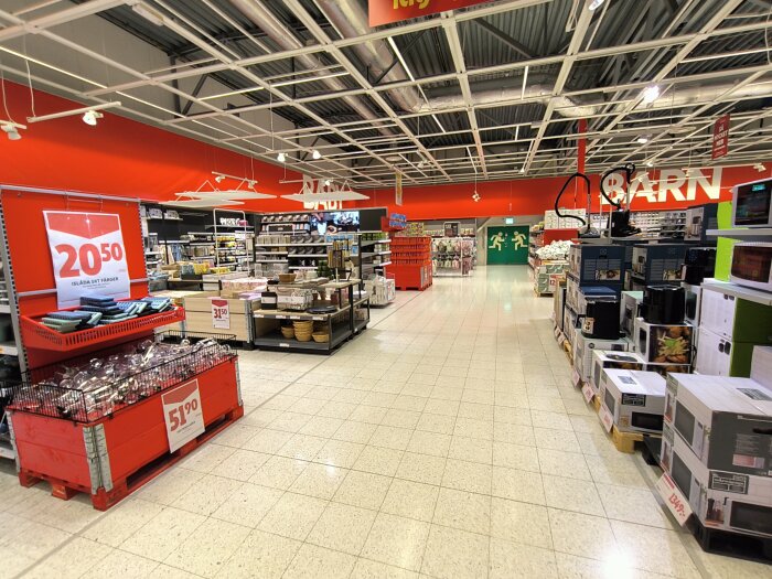 Interiör av Ica Maxi-butik med röda och vita skyltar, displayer med olika produkter och priser. Hyllor med hushållsvaror och skylt "BARN".