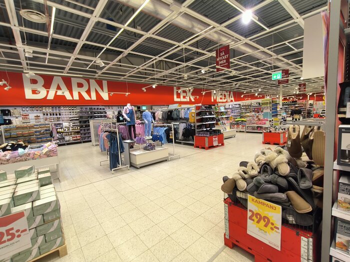 Interiör av en Ica Maxi-butik med barnsektion, kläder och leksaker. Skyltar med kategorierna "Barn", "Lek" och "Media" syns på röda bakgrunder.