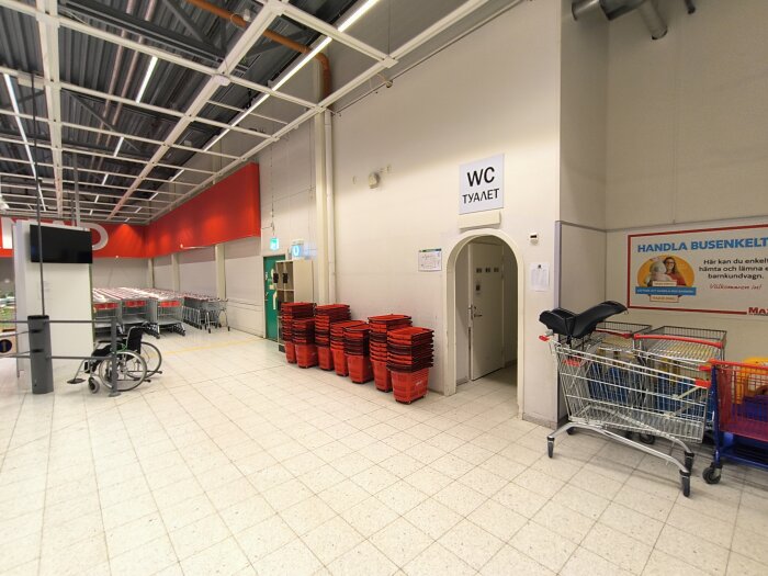 Interiör av en Ica Maxi-butik med kundvagnar och korgar vid ingång till en toalett, skyltar på svenska och ryska, samt reklamskylt om kundvagnstjänst.