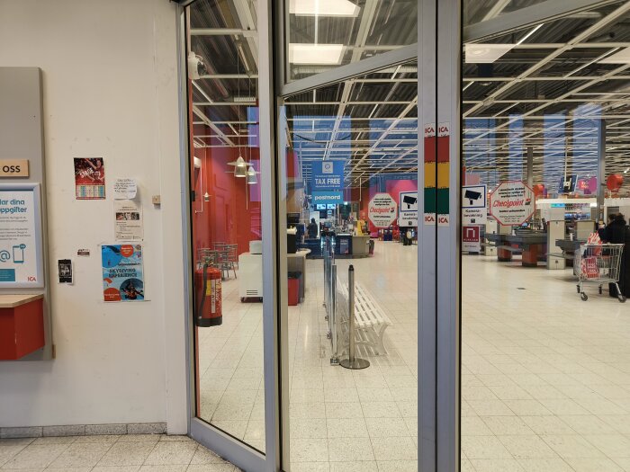 Interiör av en Ica Maxi-butik med skyltar för kameraövervakning och självskanning, ingång med glasdörr och posters på väggarna.