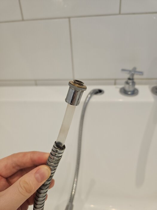 Hand håller duschslang med exponerad intern slang; synlig vit badrumsvägg och badkarskran i bakgrunden.