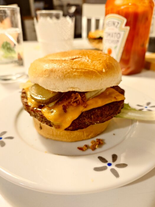 Hamburgare med ost, pickles, och sallad på vit tallrik med blomdesign. Ketchupflaska och glas i bakgrunden.