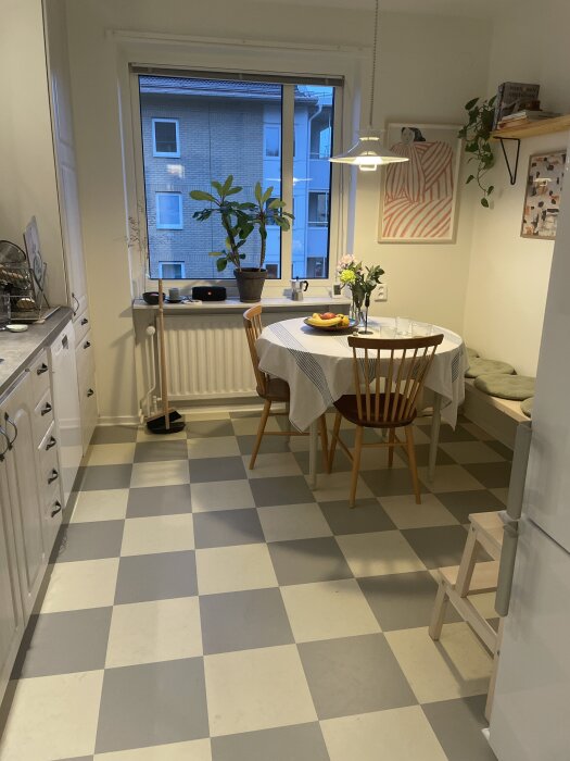 Kök med vit och grå schackrutigt golv, matbord med stolar och växter på fönsterbrädan, sett från ingången.