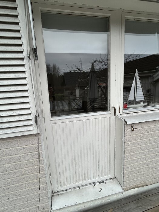 Vit altandörr från 1975 med glasruta, sedd utifrån, omges av ljus tegelvägg och träpanel.