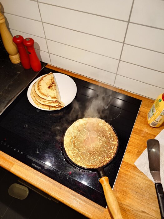 Stekpanna med pannkaka på en spis, rykande pannkaka, tallrik med färdiga pannkakor bredvid.