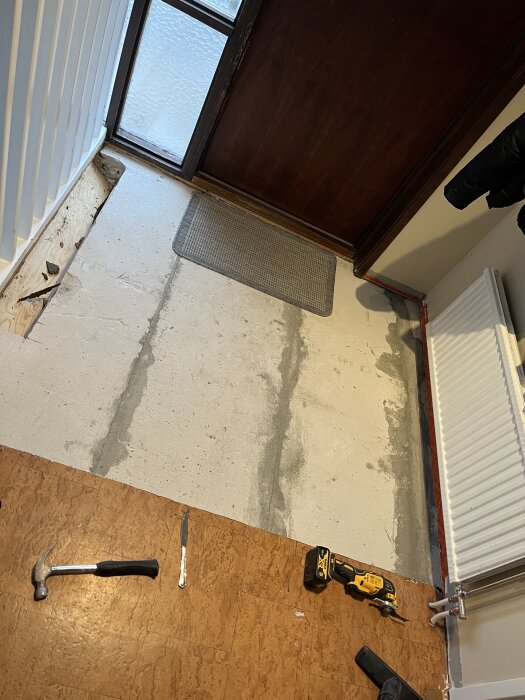Renovering av hallgolv med verktyg som hammare, kniv och slipmaskin på korkmatta. Dörr och radiator syns i bakgrunden.