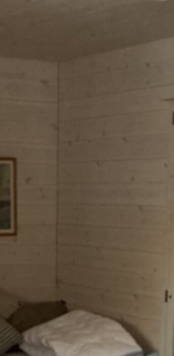 Et hjørne i et nyligt bygget attefallshus med trælister på væg og loft. Synlige små sprækker mellem panelerne. En seng er delvist synlig i nederste venstre hjørne.