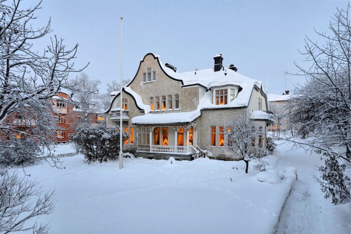 Snötäckt villa med karaktäristiska takkonstruktioner, omgiven av snötyngda träd; fönstren lyser varmt i skymningen.