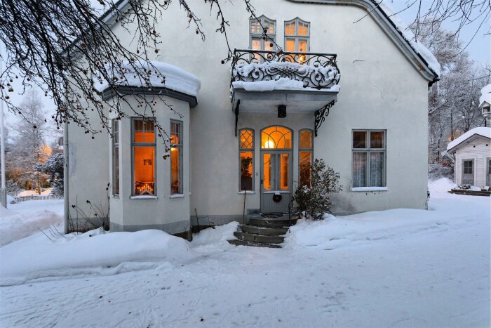 Snötäckt sekelskifteshus med varm belysning, vackert järnräcke vid balkongen och frostiga detaljer.