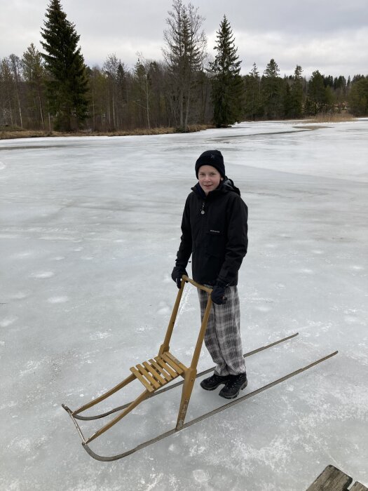 Person på frusen sjö med spark, vintermössa och jacka, omgiven av skog.