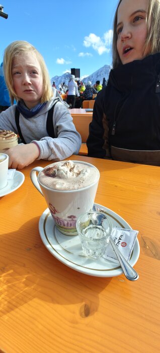 Barn sitter vid träbord utomhus med varm choklad med grädde och ett glas, berg i bakgrunden.