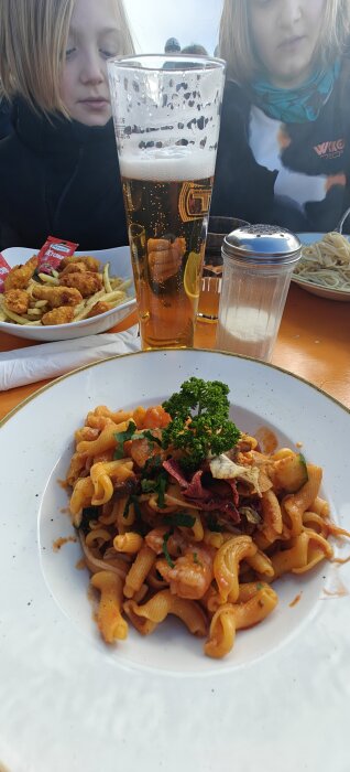 Tallrik med pasta med sås, dekorerad med persilja, tillsammans med två glas dryck, en skål pommes och nuggets, samt en saltströare på träbord.
