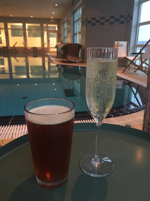 Två drycker, en öl och ett glas bubbel, placerade på ett bord vid en inomhuspool med vilstolar och dämpad belysning.