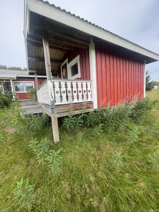 Röd mindre stuga med vita detaljer och veranda, stående på betongfundament i gräsbevuxen slänt.