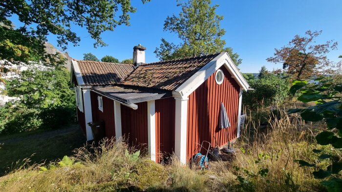 Rött trähus med små fönster och tegeltak, omgiven av träd och en gräsyta, under en klarblå himmel.