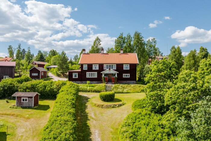 Stort trähus med röda tak och omgivande trädgård i lantlig miljö.