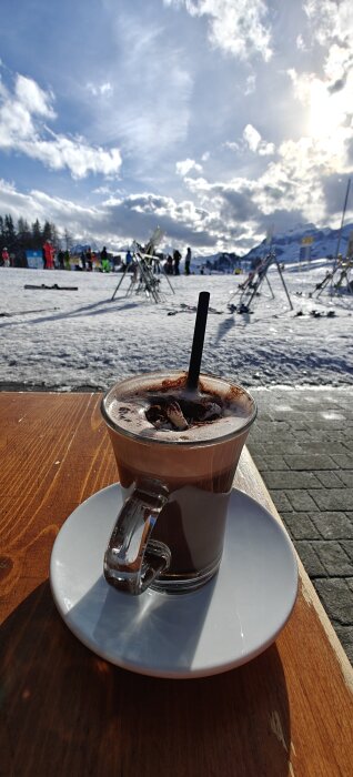 Varm choklad med marshmallows och ett sugrör på ett bord utomhus i ett snötäckt, soligt vinterlandskap.