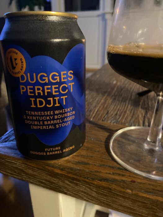 Burk med Dugges Perfect Idjit, en imperial stout lagrad på whisky och bourbonfat, bredvid ett glas på träbord.