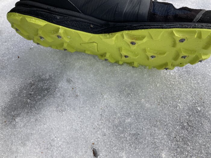 Skosula med dubbar för is och snö, visar en grön sula med metallspikar, fotad på isigt underlag.