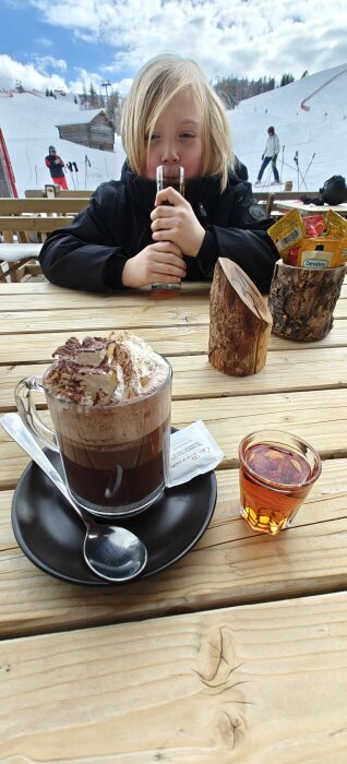 En kopp choklad med grädde och ett glas amaretto på ett bord, med ett barn som dricker i bakgrunden. Snöig skidbacke syns.