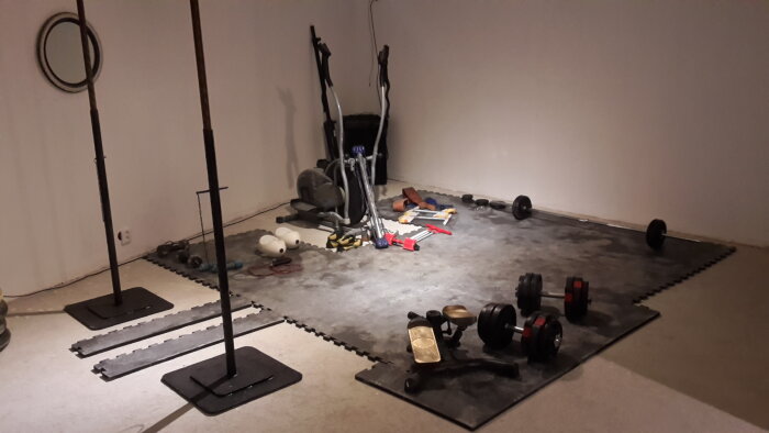 Panorama av källare med gymutrustning och verktyg på golvet, där betong delvis tagits bort för att undersöka dräneringsproblem.