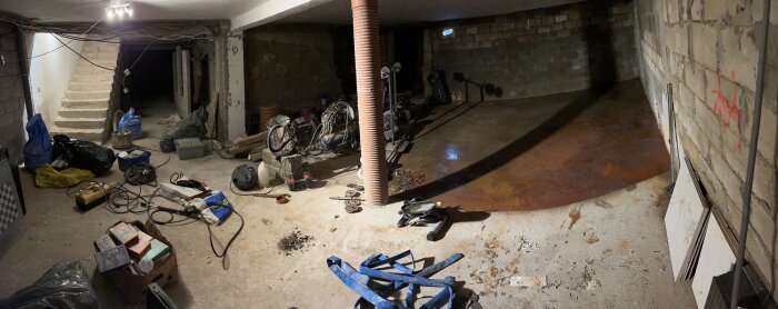Panoramabild av källare med betonggolv, byggmaterial, dräneringsrör, och en fuktfläck nära väggen. Stökig miljö med verktyg och trappor.