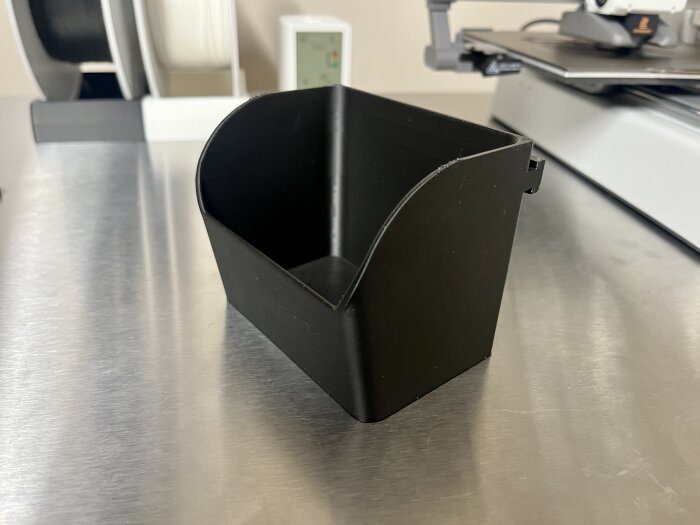 Svart 3D-utskriven plastlåda designad för att passa i en verktygsskena, placerad på en metallbänk bredvid en 3D-skrivare.