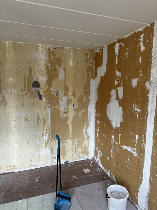 Ett gästrum under renovering med gamla tapeter borttagna, synliga blåbetongväggar och sprickor. En skurborste och hink syns på golvet.