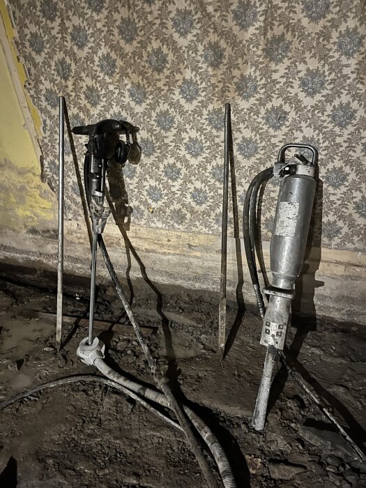 Verktyg för rivning och håltagning, inklusive en borrmaskin och hydraulisk spräckare, på en byggarbetsplats med mönstrad vägghängd bakgrund.