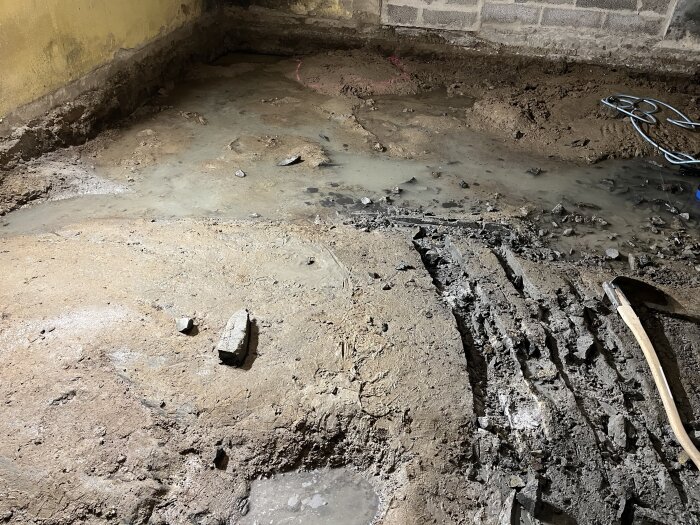En bild av en grusig golvyta i ett rum under renovering, med synliga skrapmärken och en pöl med vatten. En spade ligger på marken.