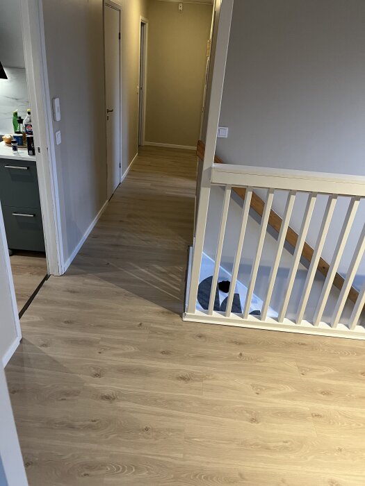 Korridor med ljust trägolv, vita väggar och trappa med räcke. Kök syns delvis till vänster.