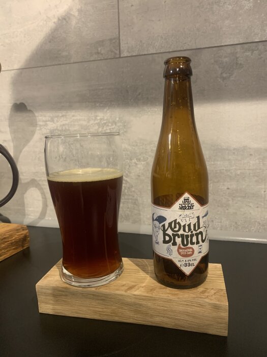 Flaska och glas med belgisk öl, märkning "Oud Bruin" på flaskan, på en träbricka mot bakgrund av kakelvägg.