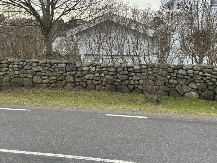 En låg stenmur byggd av ojämna naturstenar, omgiven av träd och buskar, med ett hus i bakgrunden.