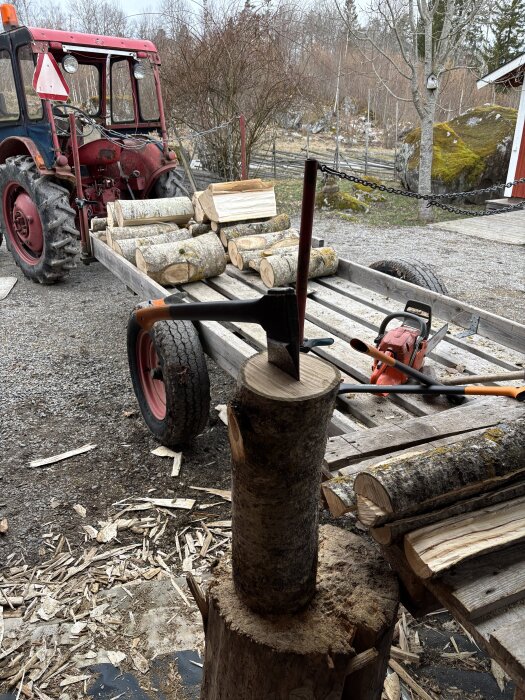 Traktor med vedkärra fylld med vedträn, yxa och motorsåg i förgrunden, omgivande gårdslandskap.