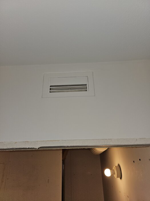 Ventilgaller i taket, rektangulärt med horisontella spjäll, ovanför en enkel glödlampa på en vägg.