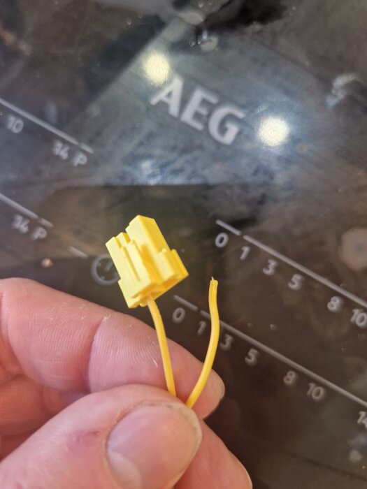 Gul trasig sensorplugg med två avklippta kablar, hålls mellan fingrarna framför baksidan av en AEG-kylskåpstermometer.
