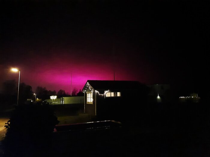 Natthimmel upplyst av rosa ljus från växthus i bakgrunden, med ett hus och gatubelysning i förgrunden.